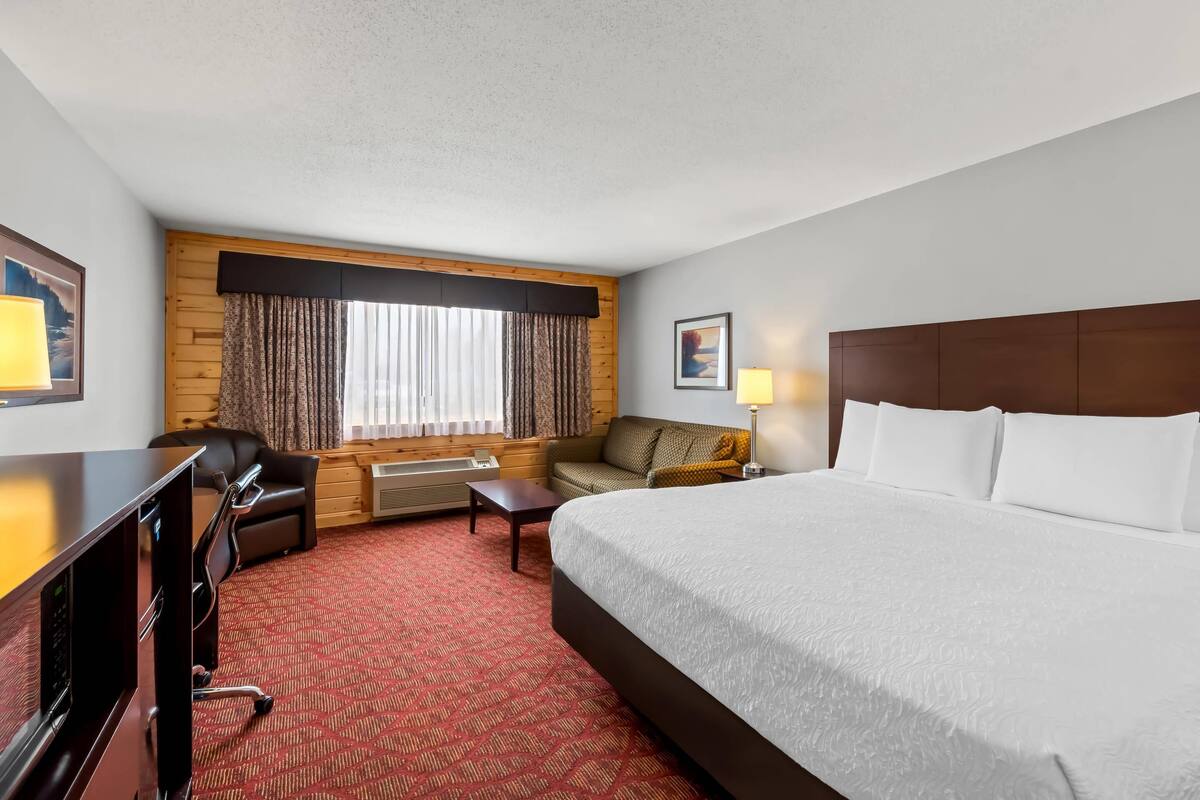A hotel room with a large bed, a desk, a chair, a sofa, a window with curtains, and a painting on the wall.