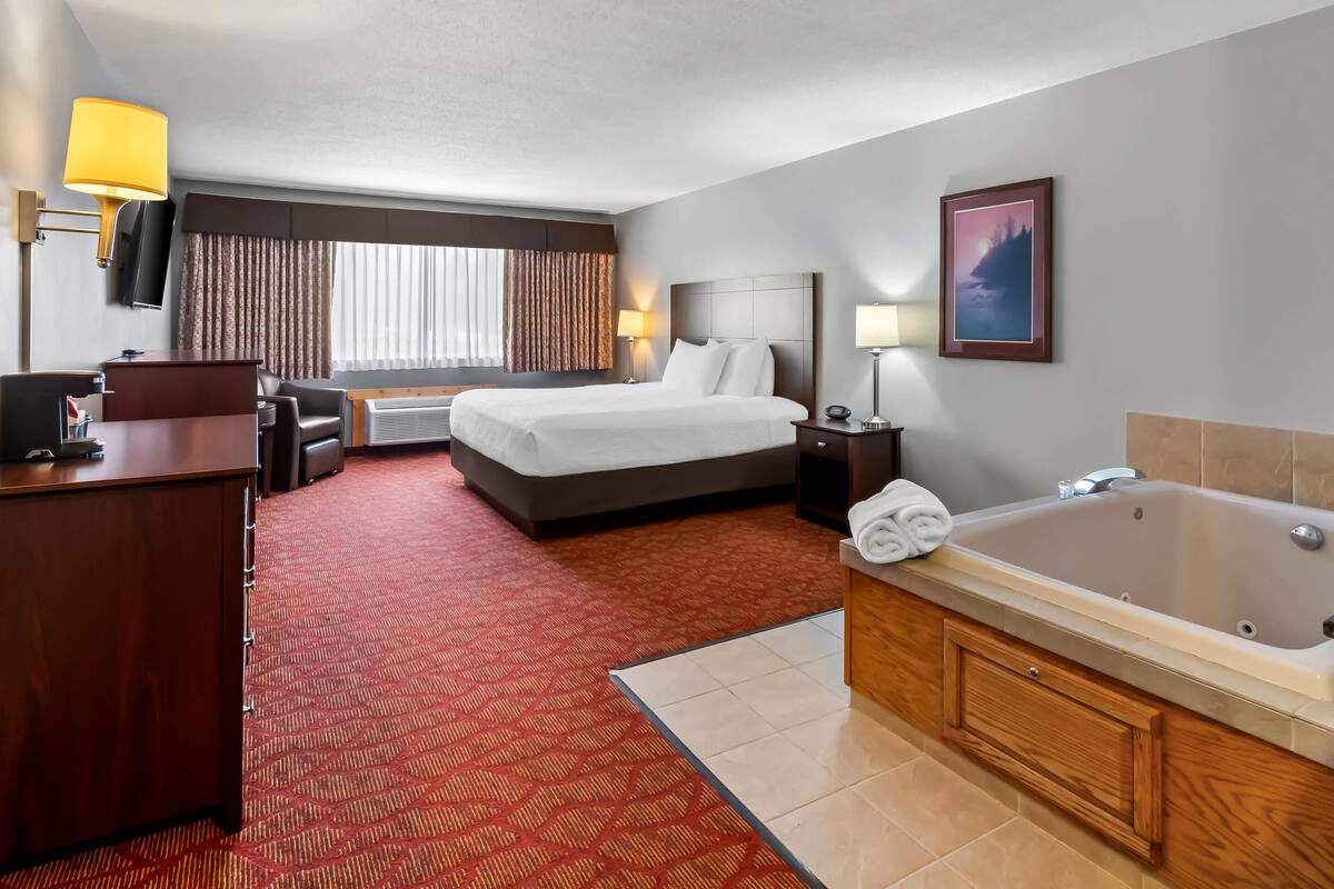 A hotel room with a large bed, a desk, a desk lamp, a TV, a bathroom with a bathtub, and a window with curtains.