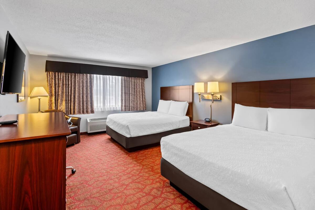 A hotel room with two beds, a desk, a TV, and a window with curtains.