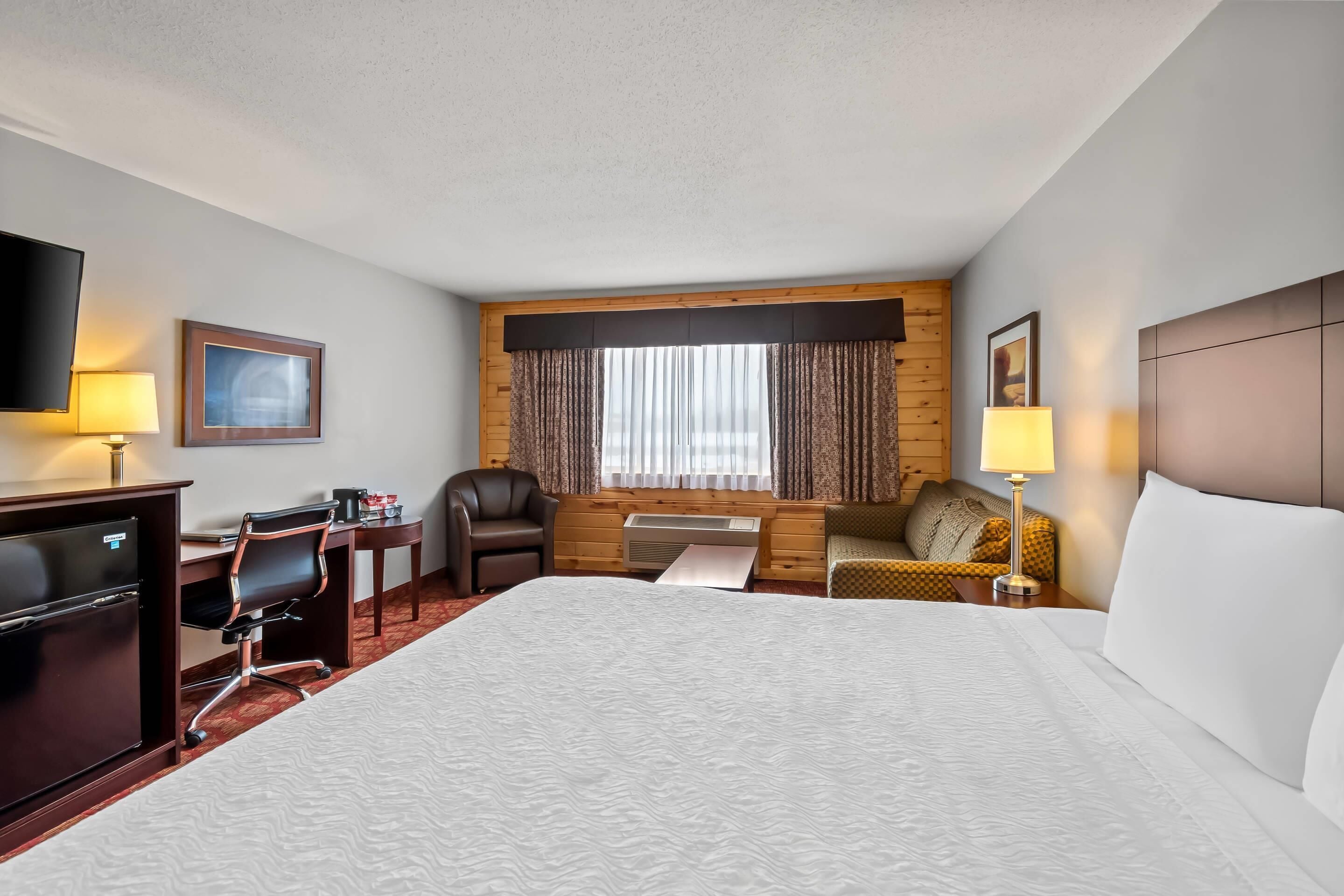 A hotel room with a large bed, a desk with a computer, a chair, a sofa, a TV, and a window with curtains.