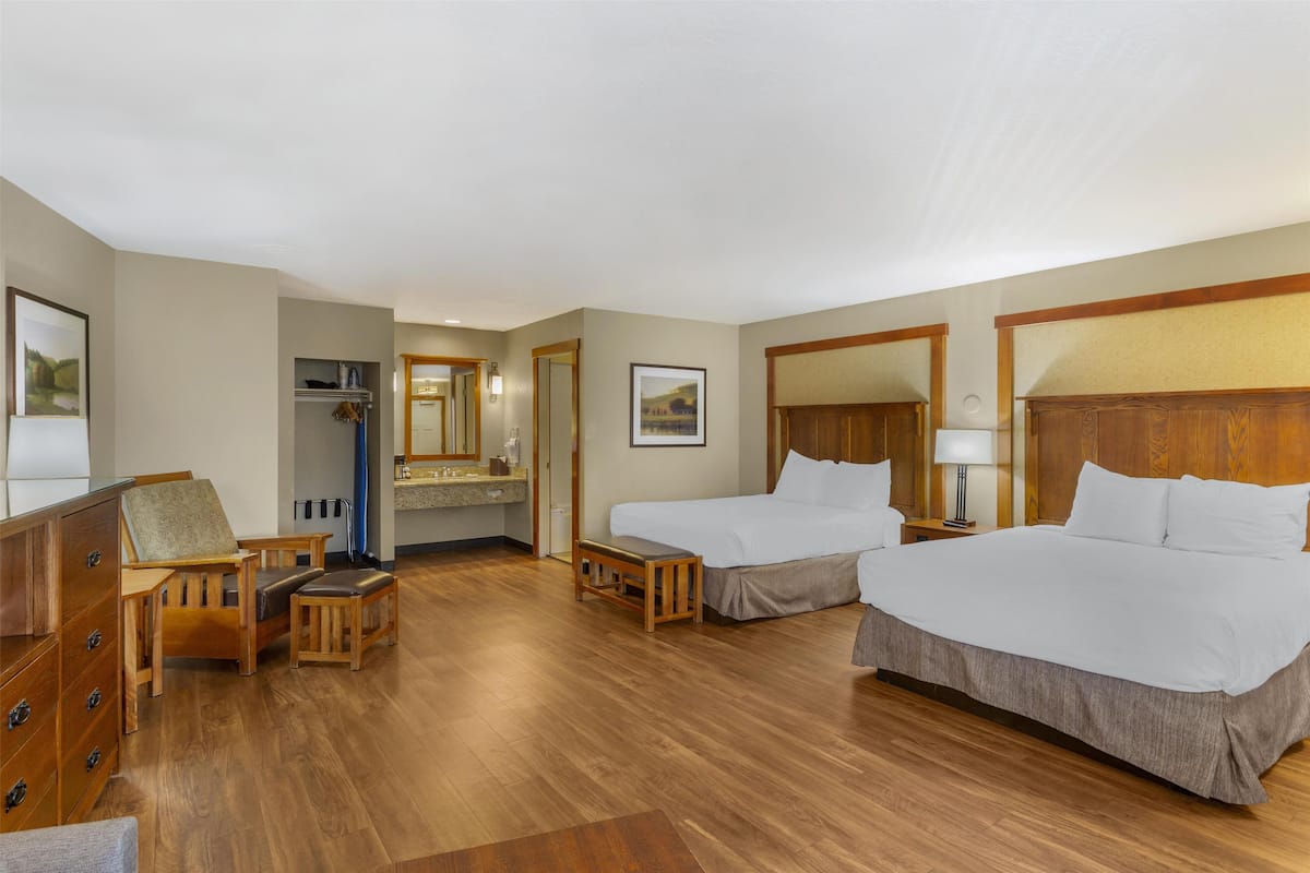 A hotel room with two beds, a wooden dresser, a desk, a chair, a small table, and a bathroom with a mirror and sink.