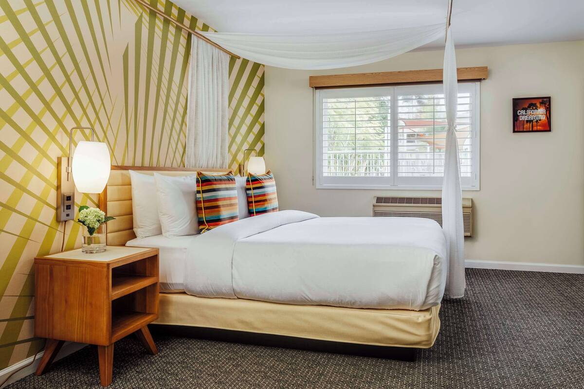 A hotel room with a bed, a bedside table with a lamp, a window with blinds, and a wall with a striped pattern.