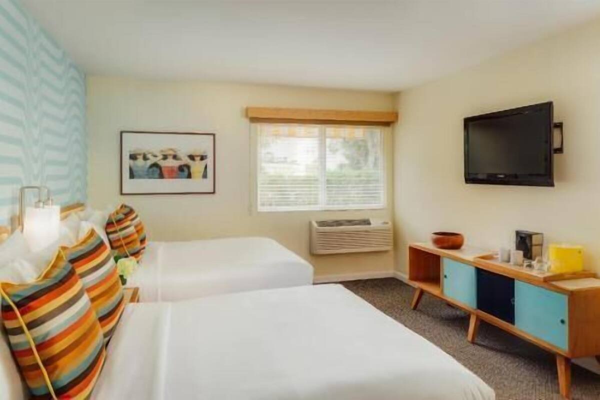 A hotel room with two beds, a television, a window with blinds, a framed picture on the wall, and a small cabinet with a bowl and cups.