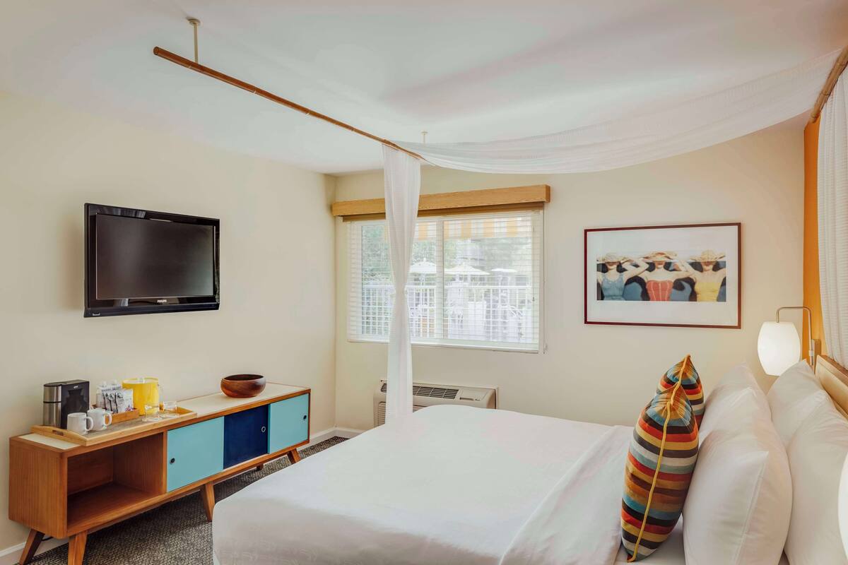 A hotel room with a bed, a TV, a window with blinds, a picture on the wall, and a bedside table with a lamp.