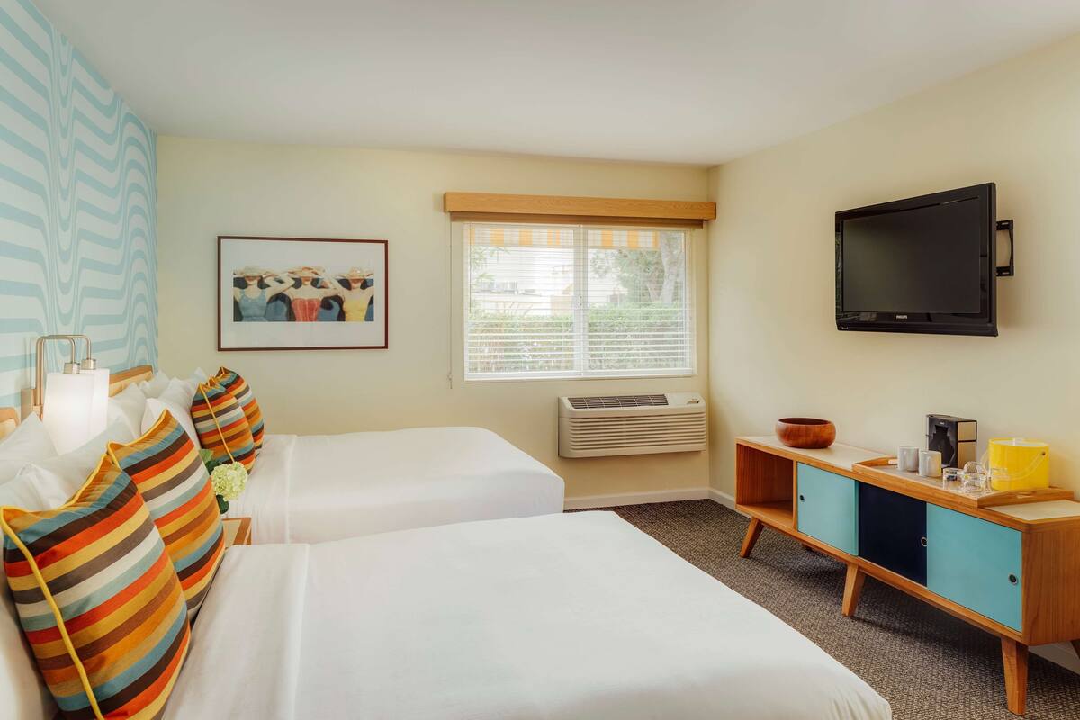 A hotel room with two beds, a television, a window with blinds, a picture on the wall, and a wooden cabinet with various items.