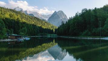 Ein stiller See mit einer Holzsteganlage und einem kleinen Gebäude, umgeben von dichtem Wald und Bergen im Hintergrund.