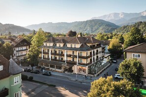 A multi-story hotel with a balcony on the second floor, surrounded by trees and a mountainous backdrop.