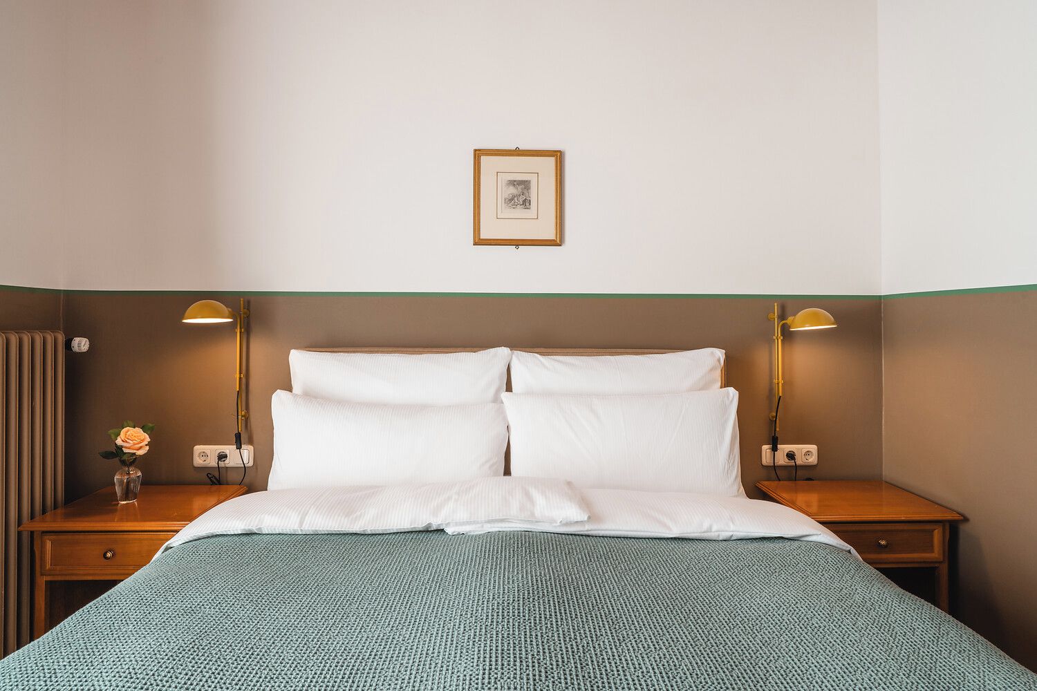 Une chambre d’hôtel avec un lit, deux tables de chevet, une petite table, une lampe, un tableau au mur et un vase avec une fleur.