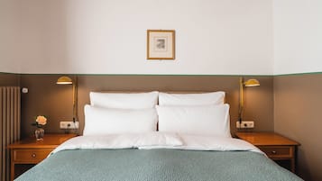 Une chambre d’hôtel avec un lit, deux tables de chevet, une petite table, une lampe, un tableau au mur et un vase avec une fleur.
