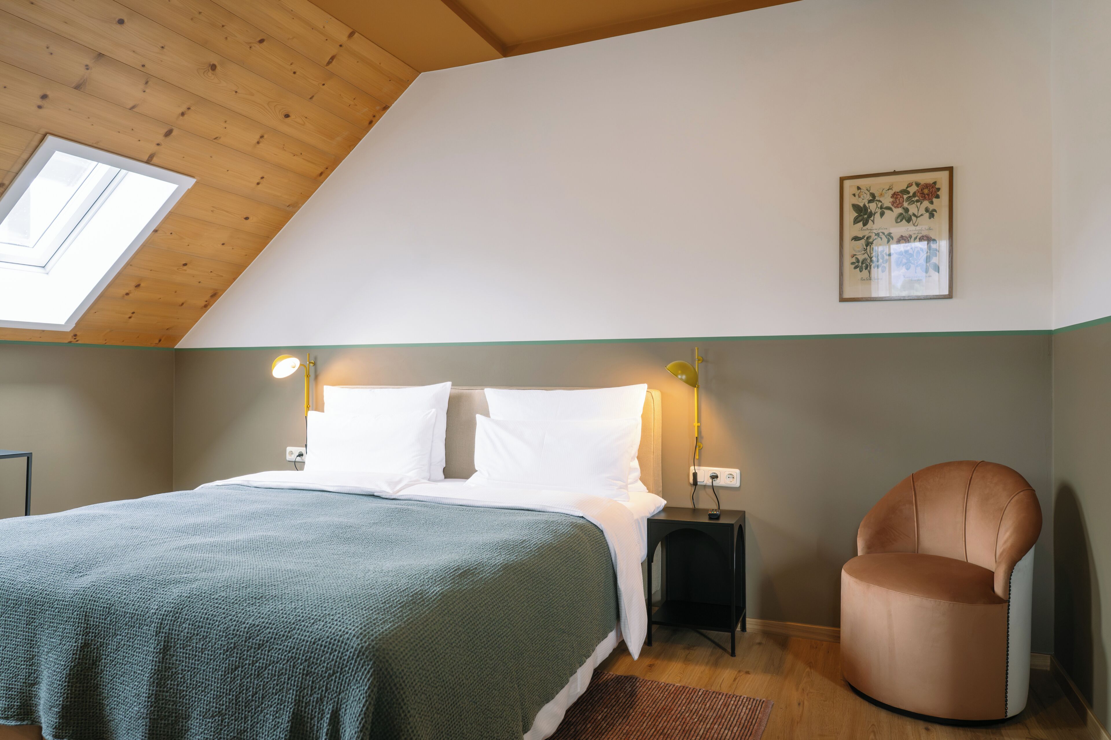 A bedroom with a sloped ceiling, a bed with white bedding, a green throw, a side table, a chair, and a framed floral artwork on the wall.