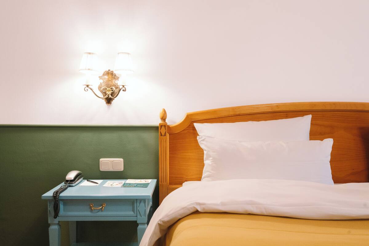 Un lit bien fait, avec du linge de lit blanc, une tête de lit en bois et une table de chevet sur laquelle se trouvent un téléphone et des papiers.