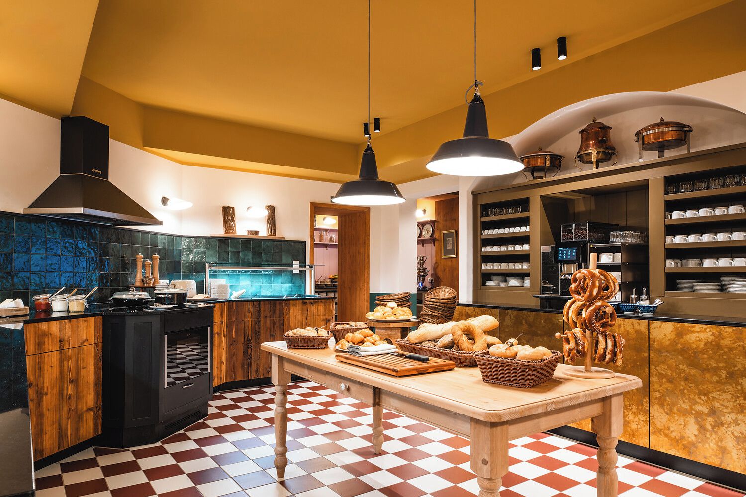 Une cuisine avec un carrelage à motifs, des meubles en bois, un îlot central sur lequel est disposé du pain, et des luminaires suspendus.