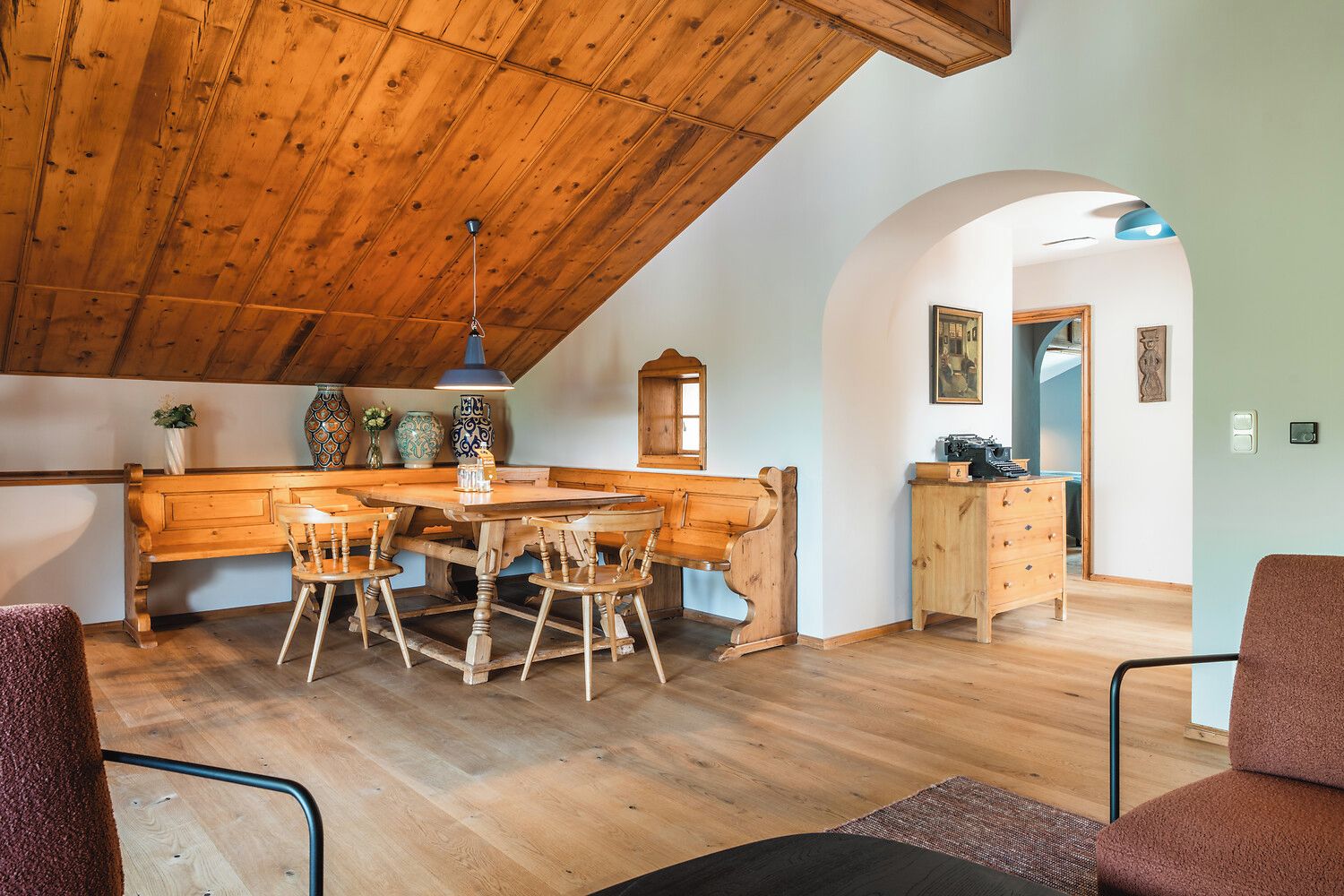 A rustic interior with a wooden dining table, chairs, and a kitchen area with wooden cabinets and a hanging light fixture.