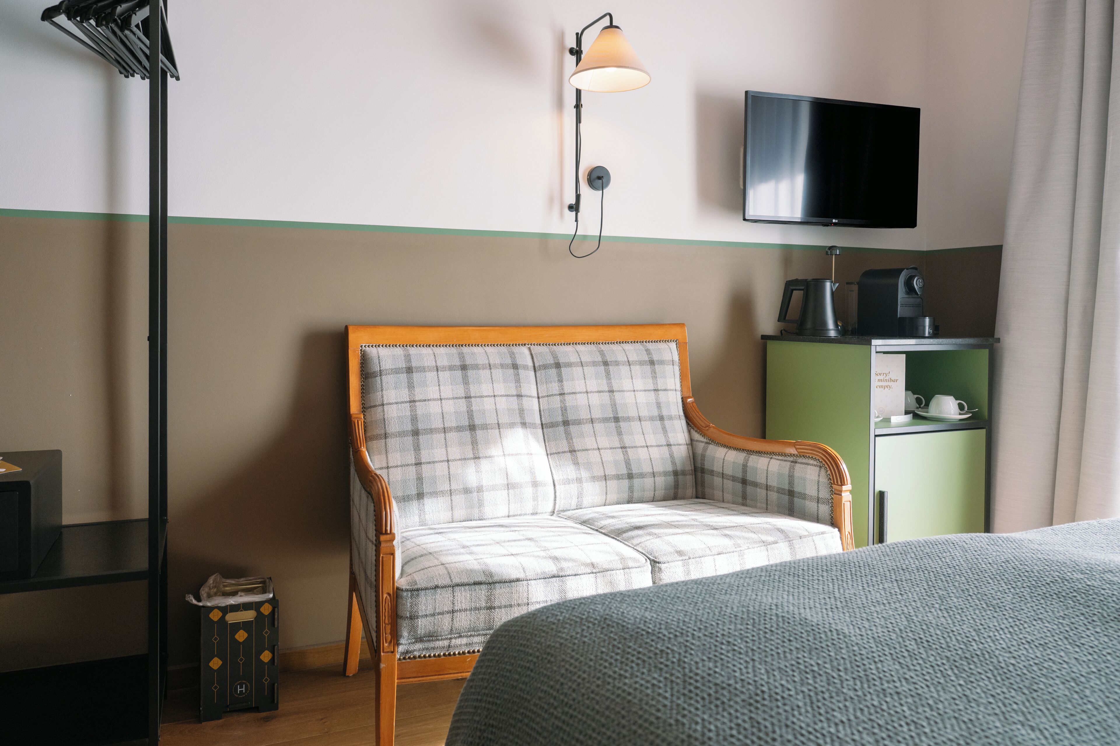 Une chambre d’hôtel avec un fauteuil à motifs écossais, un téléviseur fixé au mur, un mini-bar vert et un plancher en bois.