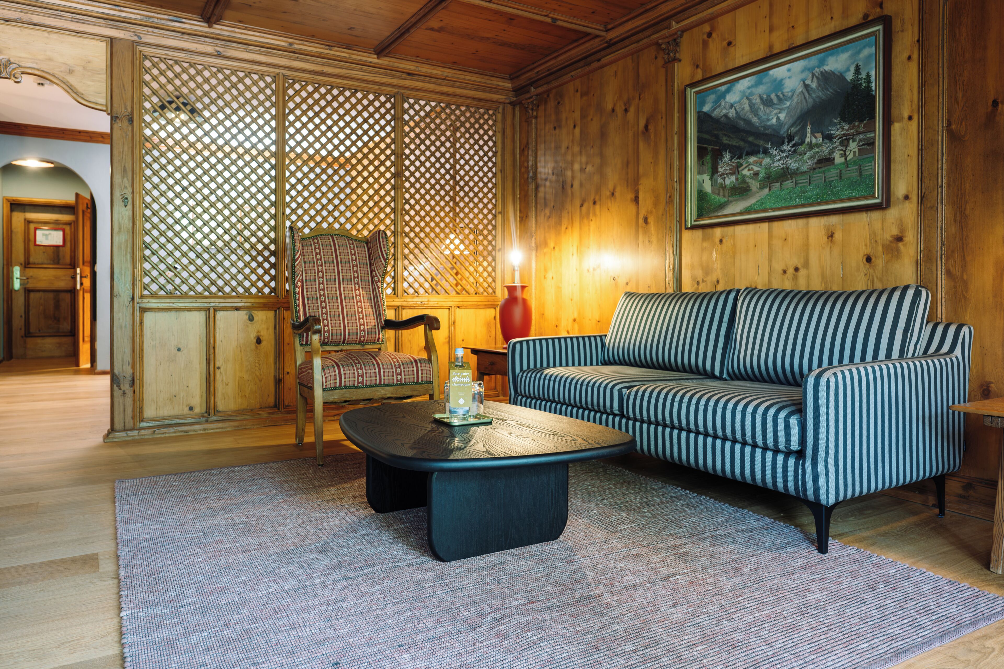 A cozy room with a wooden floor, a striped sofa, a small table with a bottle, a chair, and a painting on the wall.