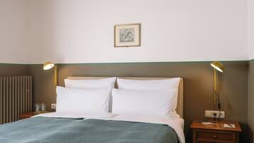 Quarto de hotel com uma cama, duas luminárias, uma mesa de cabeceira, um quadro na parede e um radiador.