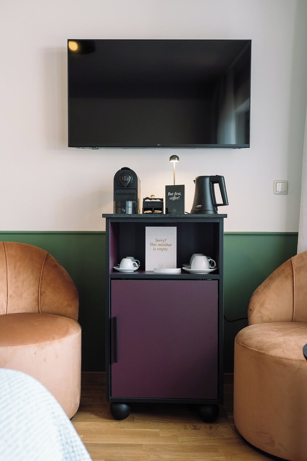 Una habitación con una televisión montada en la pared, una cafetera, un cartel que dice 'Pero primero, café' y un cartel que dice '¡Lo sentimos! Este minibar está vacío'. Hay dos sillas en la habitación.