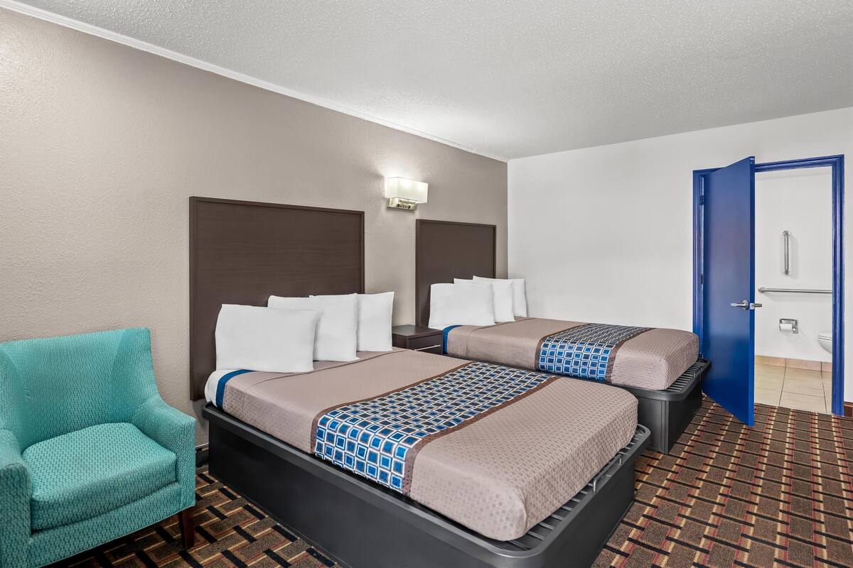 A hotel room with two beds, a teal armchair, a wall-mounted light, and a blue door.