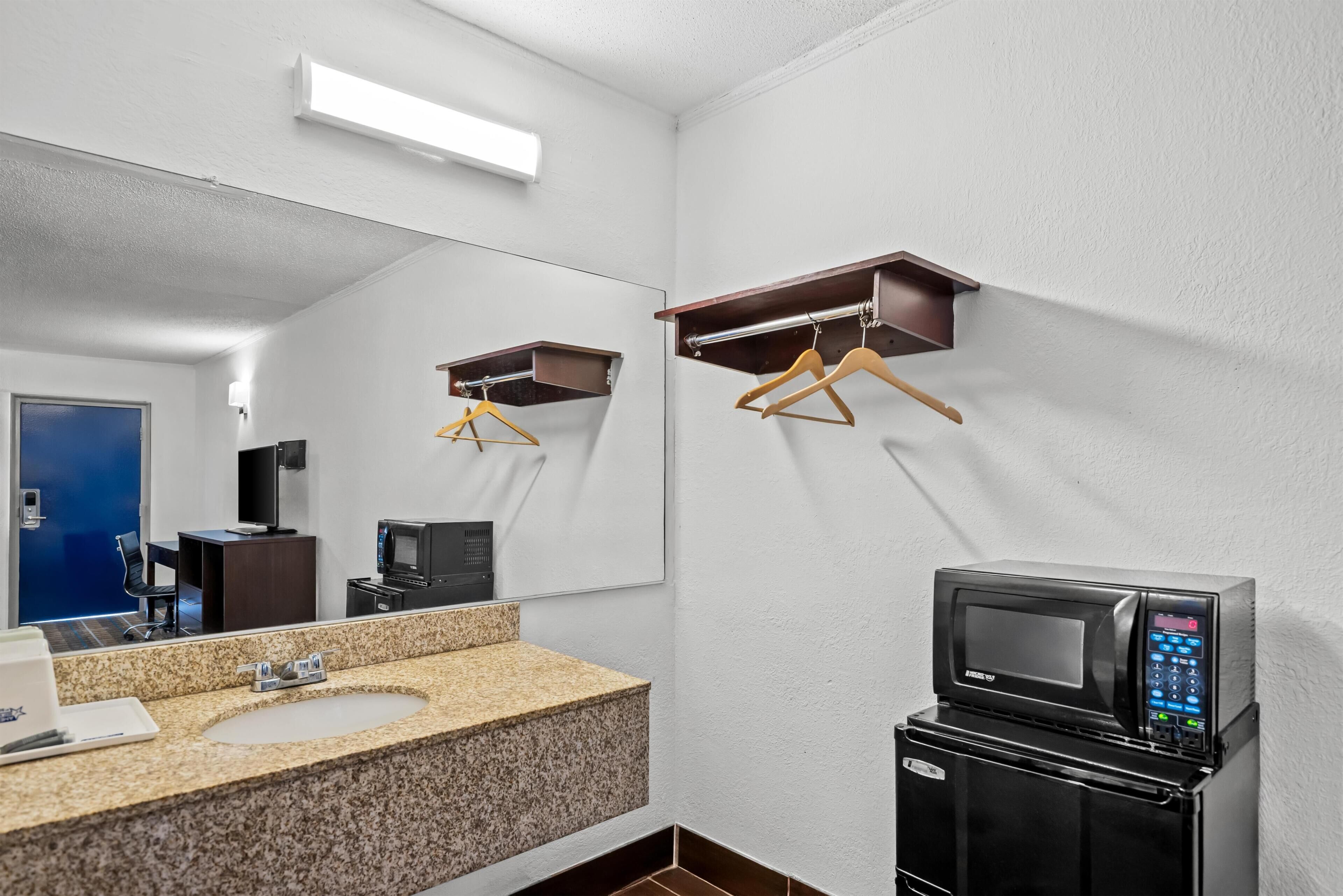 A bathroom with a granite countertop sink, a mirror, a microwave, and wooden hangers on a wall-mounted rack.