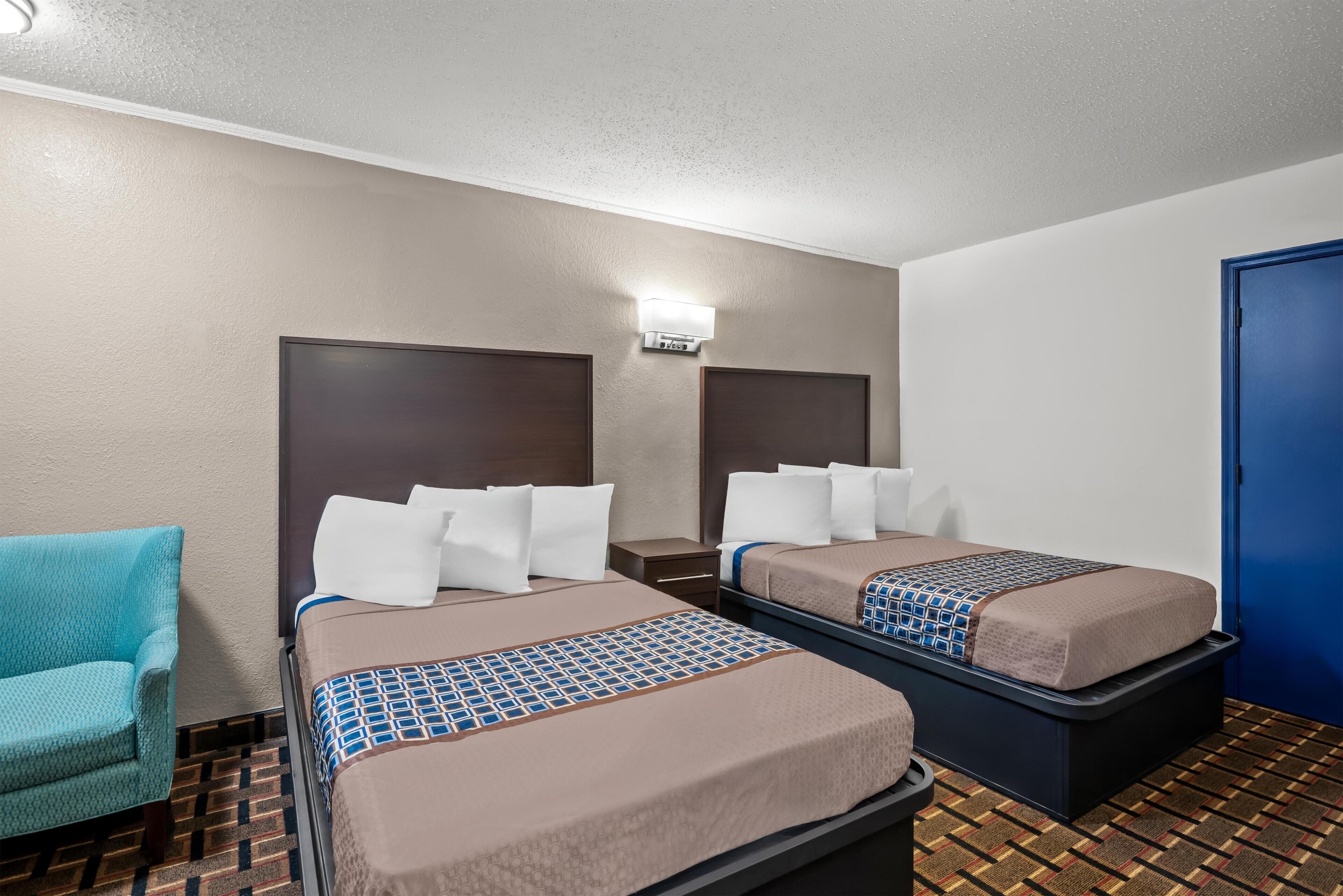A hotel room with two beds, a chair, a nightstand, and a wall-mounted light.
