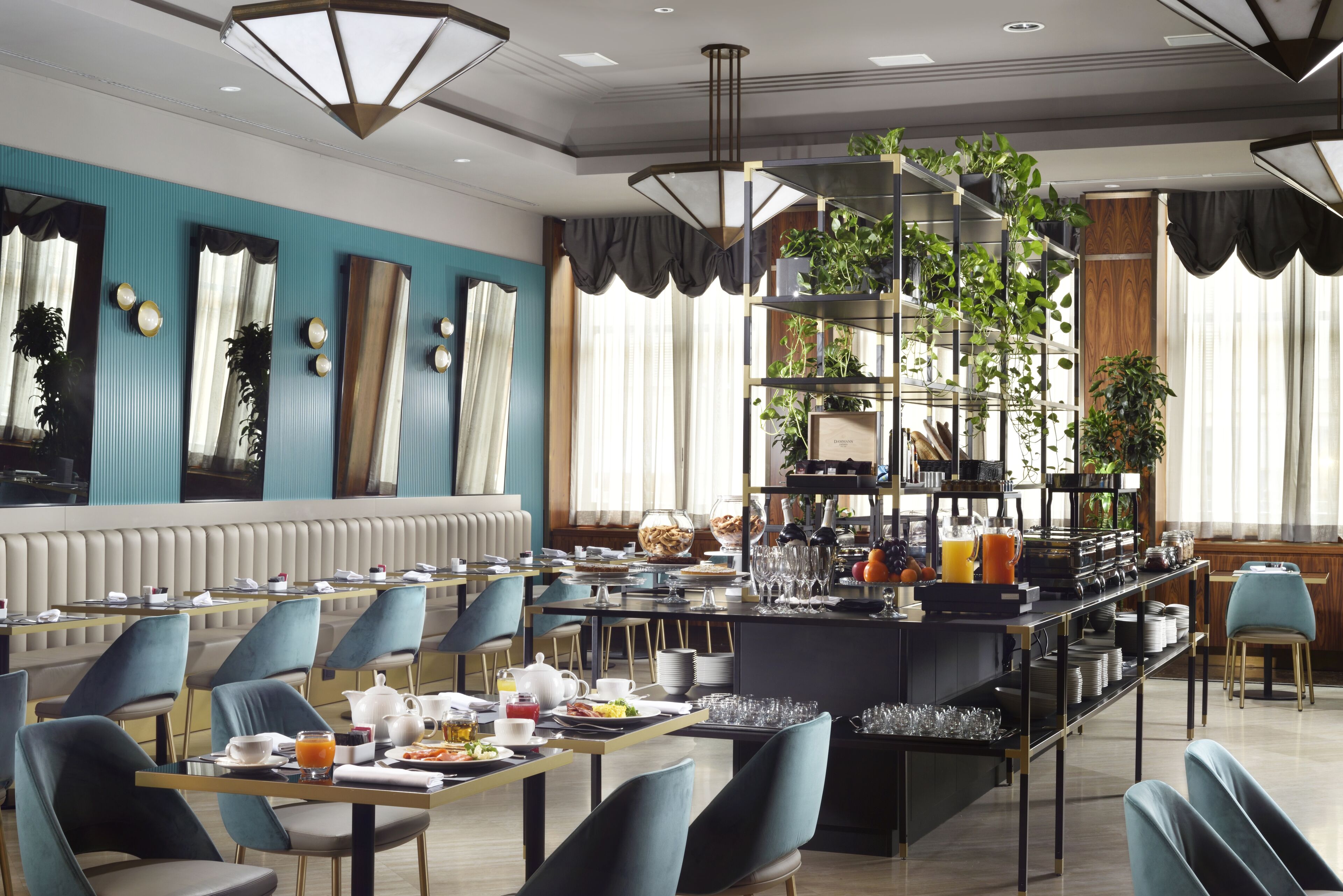 A modern dining area with a long communal table, blue chairs, and a central bar area with shelves of plants and kitchenware.