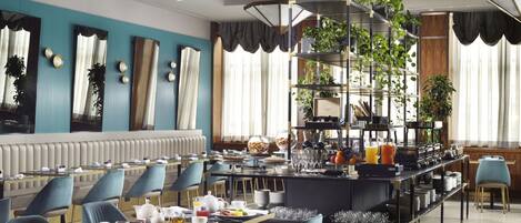 A modern dining area with a long communal table, blue chairs, and a central bar area with shelves of plants and kitchenware.