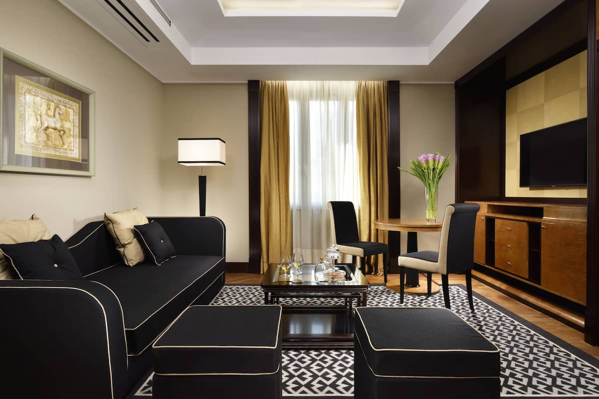 Ein modernes Hotelzimmer mit einer schwarzen Couch, einem Couchtisch, einem Fernseher und einem Essbereich mit einem Blumenstrauß.