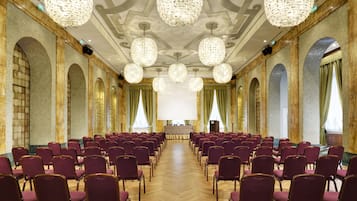 A conference room with rows of purple chairs, a large screen at the front, and ornate chandeliers.