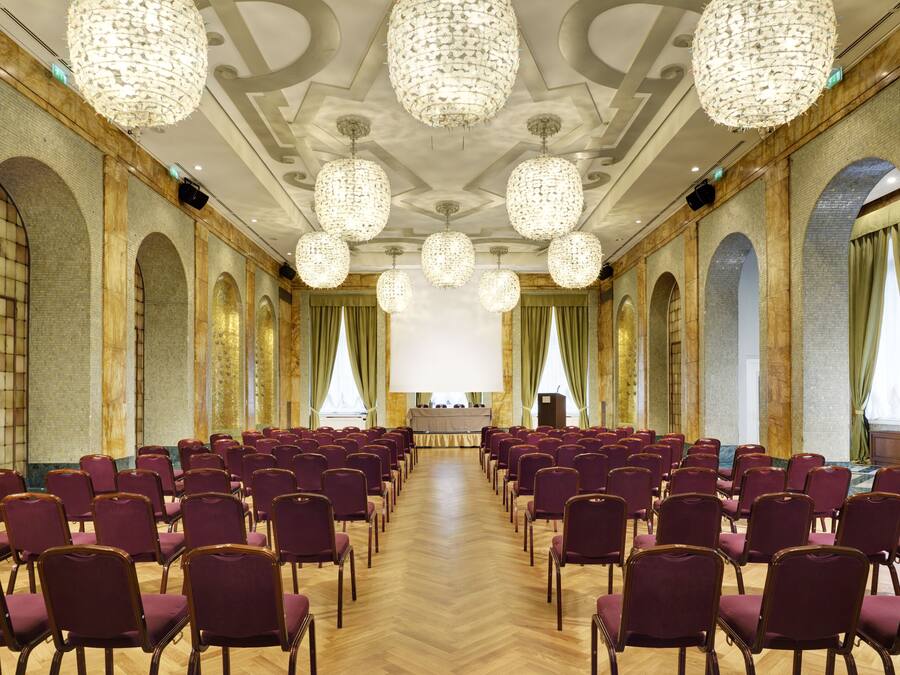 A conference room with rows of purple chairs, a large screen at the front, and ornate chandeliers.