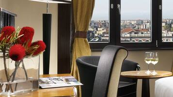 A hotel room with a bed, a chair, a table with a glass of wine, a lamp, and a vase with red flowers.