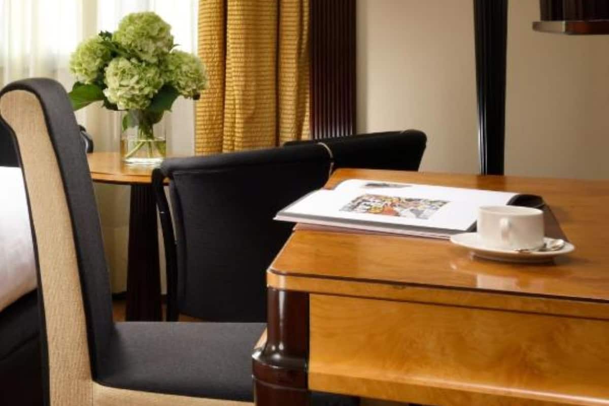 Ein Hotelzimmer mit einem Holztisch, zwei Stühlen, einer Blumenvase, einer Lampe und einem Fenster mit Vorhängen.