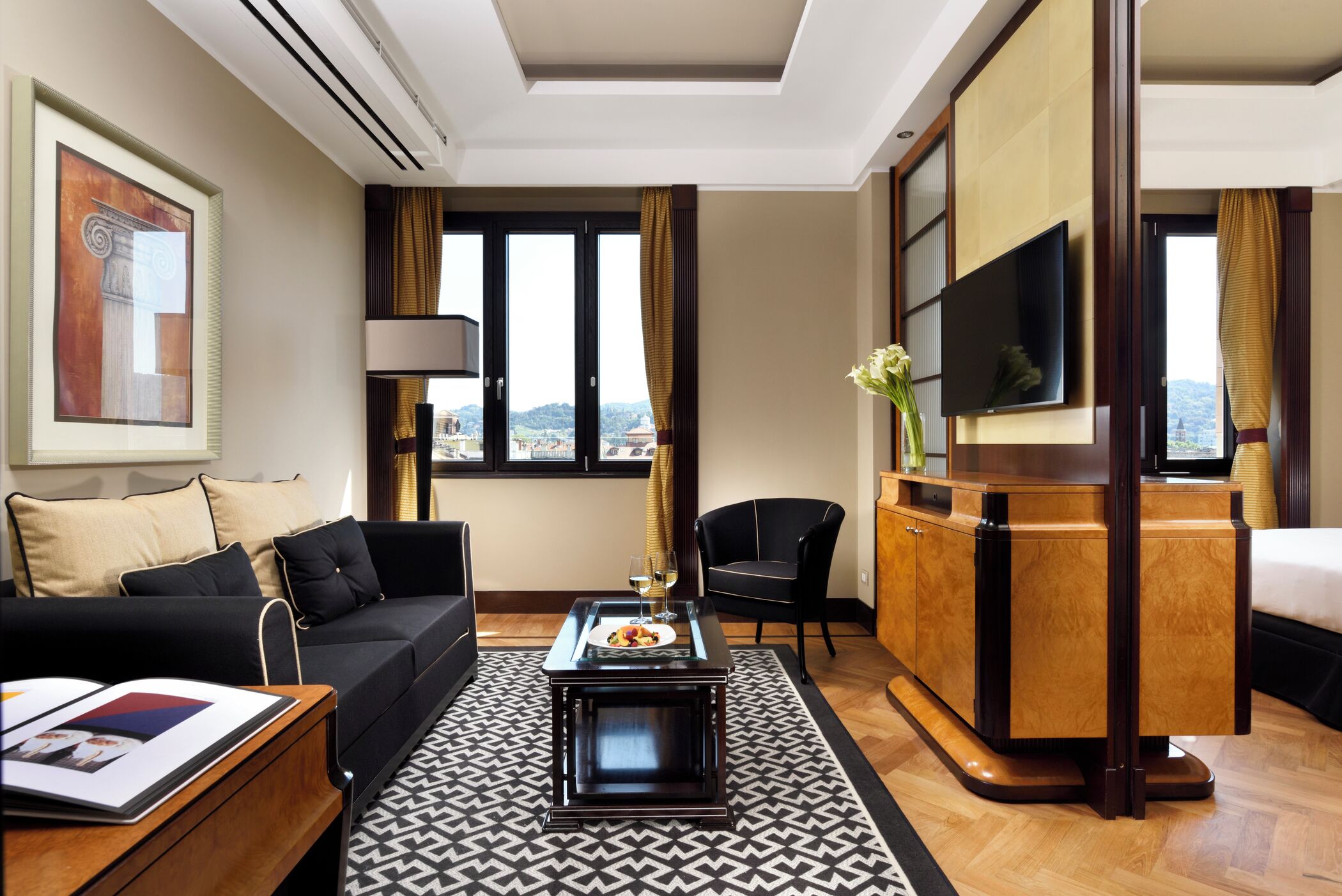 A hotel room with a sofa, a coffee table, a TV, a desk, and a view of the city.