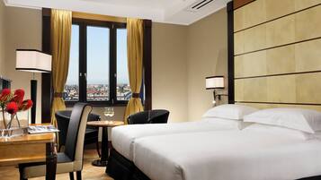 A hotel room with a bed, a desk with a lamp, a chair, and a view of the city through large windows.
