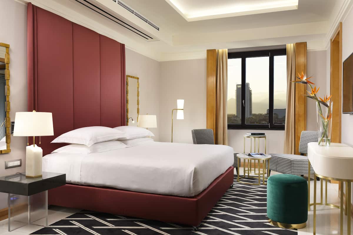 Ein Hotelzimmer mit einem großen Bett, einem Schreibtisch mit Lampe, einem Sessel, einem kleinen Tisch und Blick auf die Stadt.