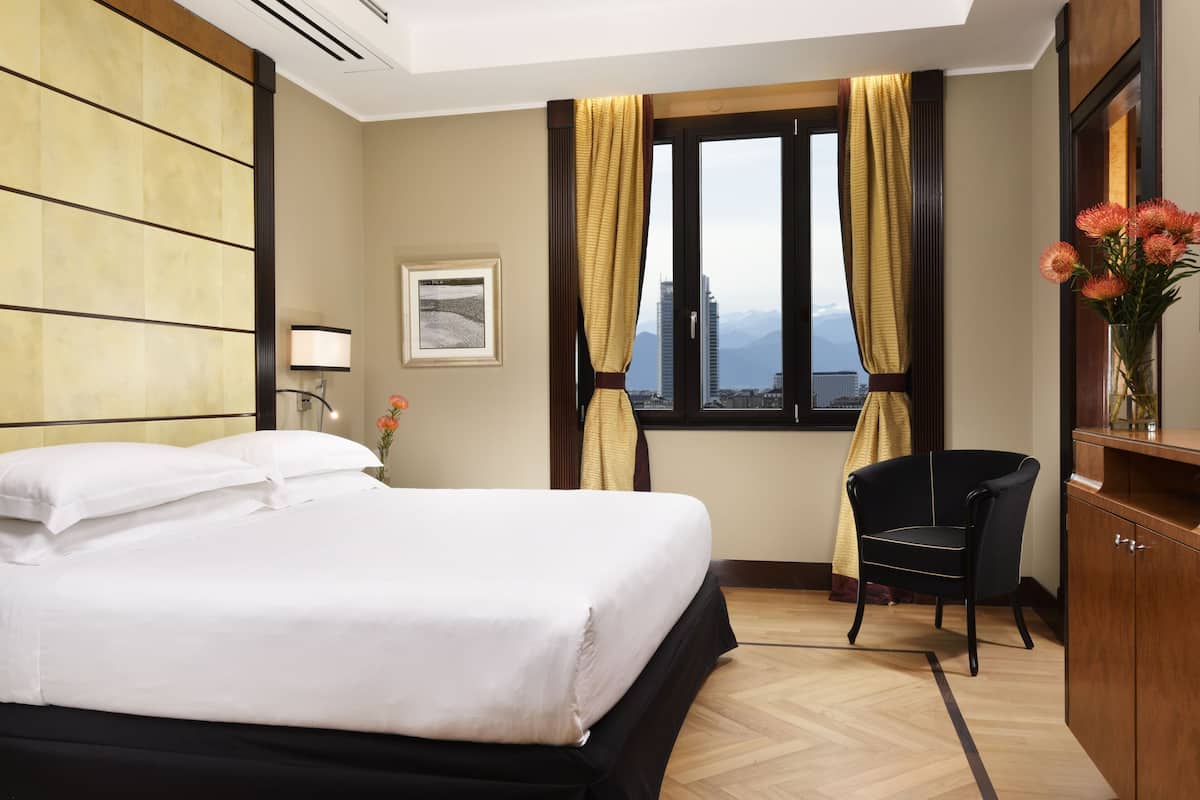 Ein Hotelzimmer mit Bett, Stuhl, Schreibtisch, einem Fenster mit Vorhängen und Blick auf die Stadt.