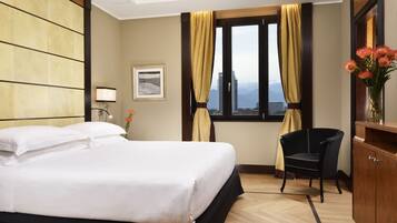 Una habitación de hotel con cama, sillón, escritorio, ventana con cortinas y vistas a la ciudad.