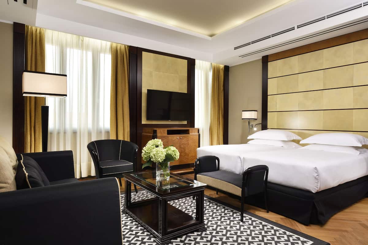 Ein Hotelzimmer mit einem Bett, zwei Stühlen, einem Couchtisch mit Blumen, einem Fernseher und einem großen Fenster mit Vorhängen.