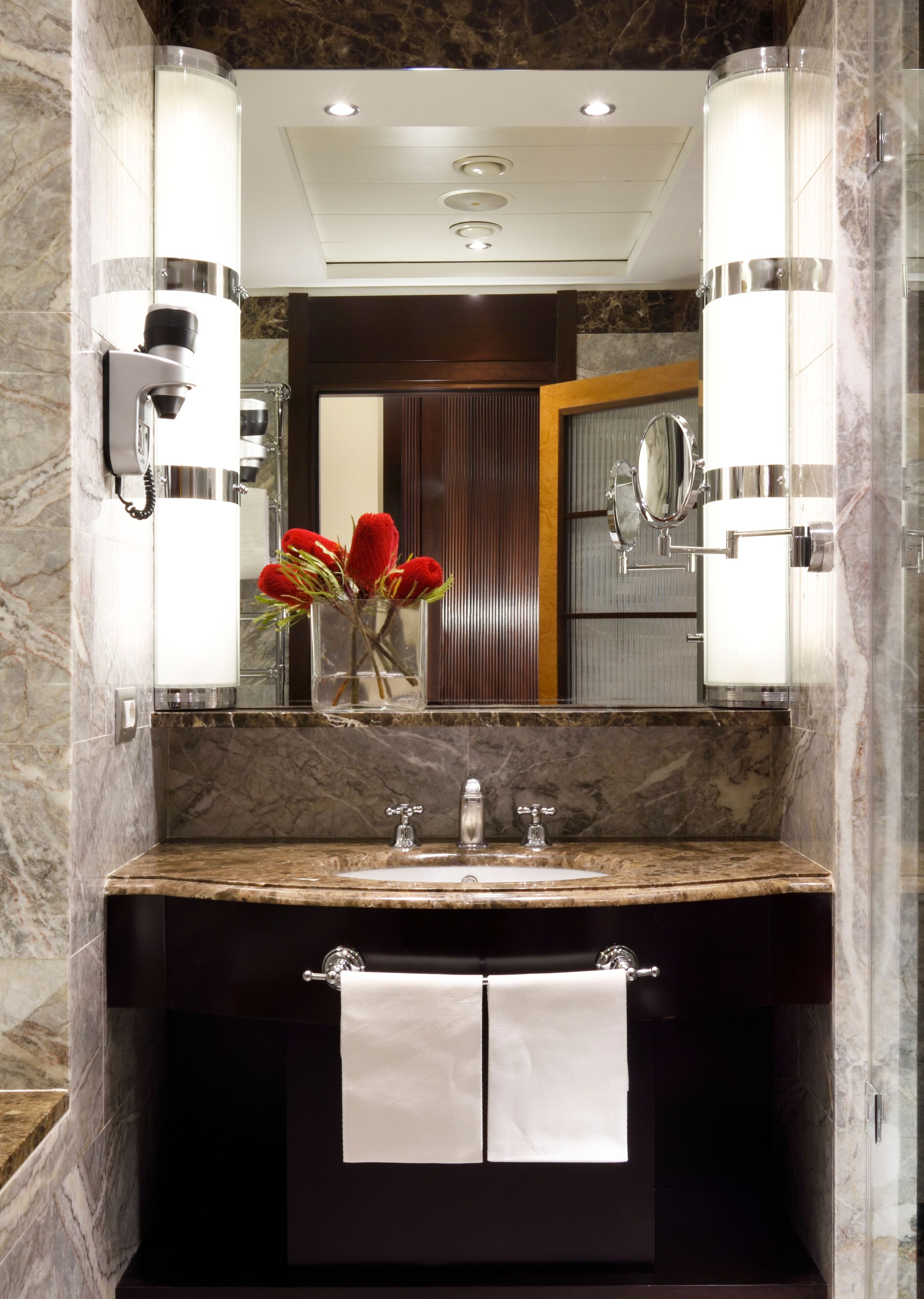 A bathroom with a marble countertop, two sinks, a mirror, and a vase with red flowers.