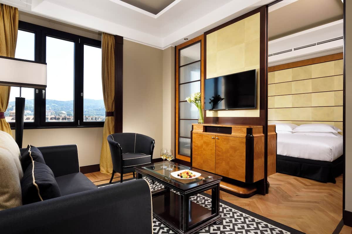 Ein Hotelzimmer mit Bett, Fernseher, Esstisch mit Obstteller, einer Couch und Blick auf die Stadt durch große Fenster.