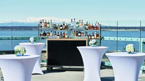A bar area with a view of a lake and mountains, featuring white tables with vases and a variety of liquor bottles. - Hilton Anchorage (Anchorage)