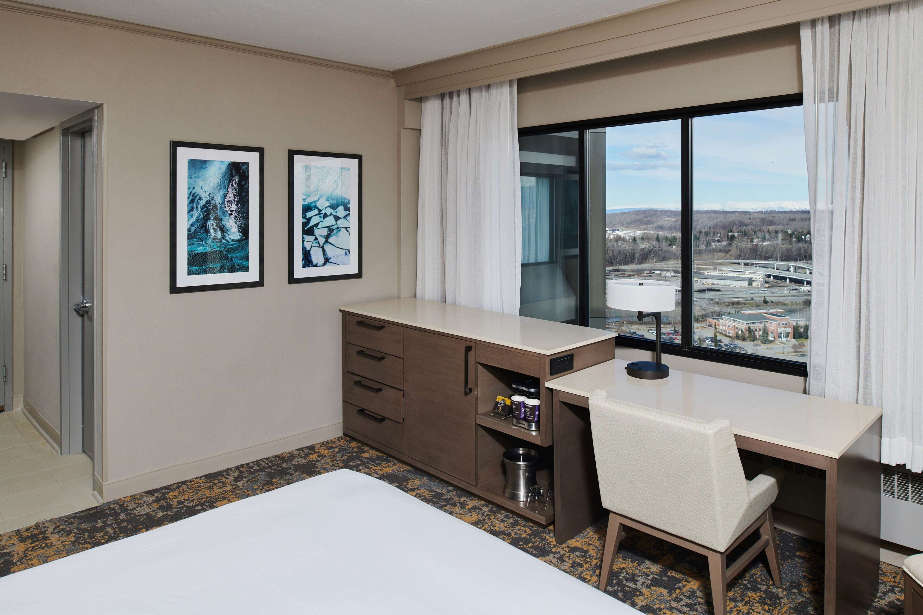 Ein Hotelzimmer mit Bett, Schreibtisch, Sessel, Minibar und einem großen Fenster mit Blick auf eine Brücke und Gebäude.