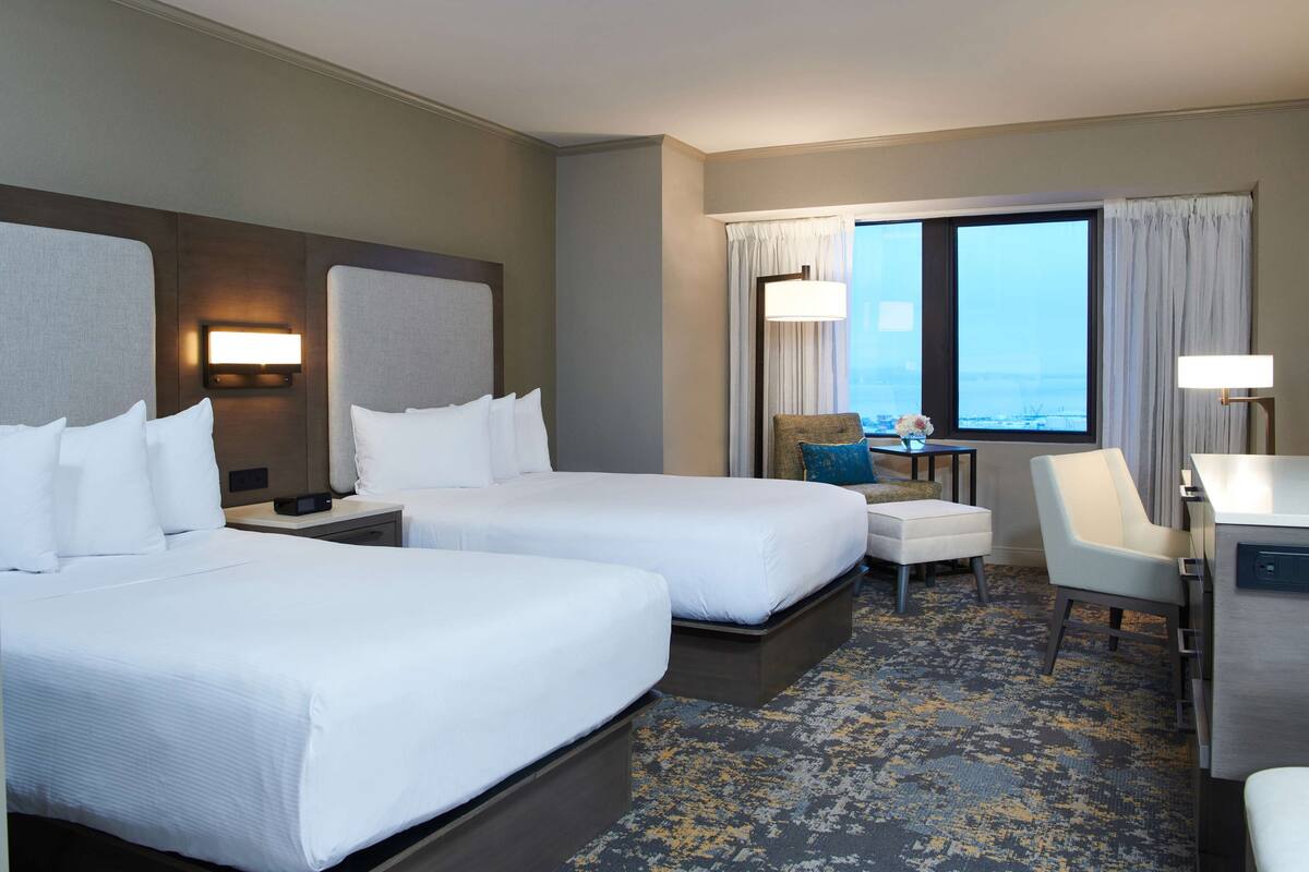 A hotel room with two beds, a desk, a chair, and a view of the city.