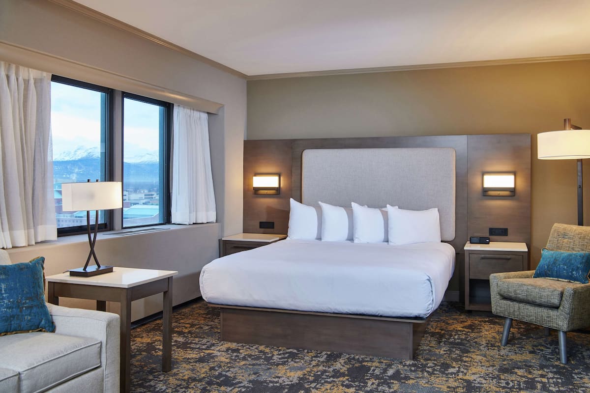A hotel room with a large bed, two bedside tables with lamps, a chair, and a view of a snowy mountain through a window.