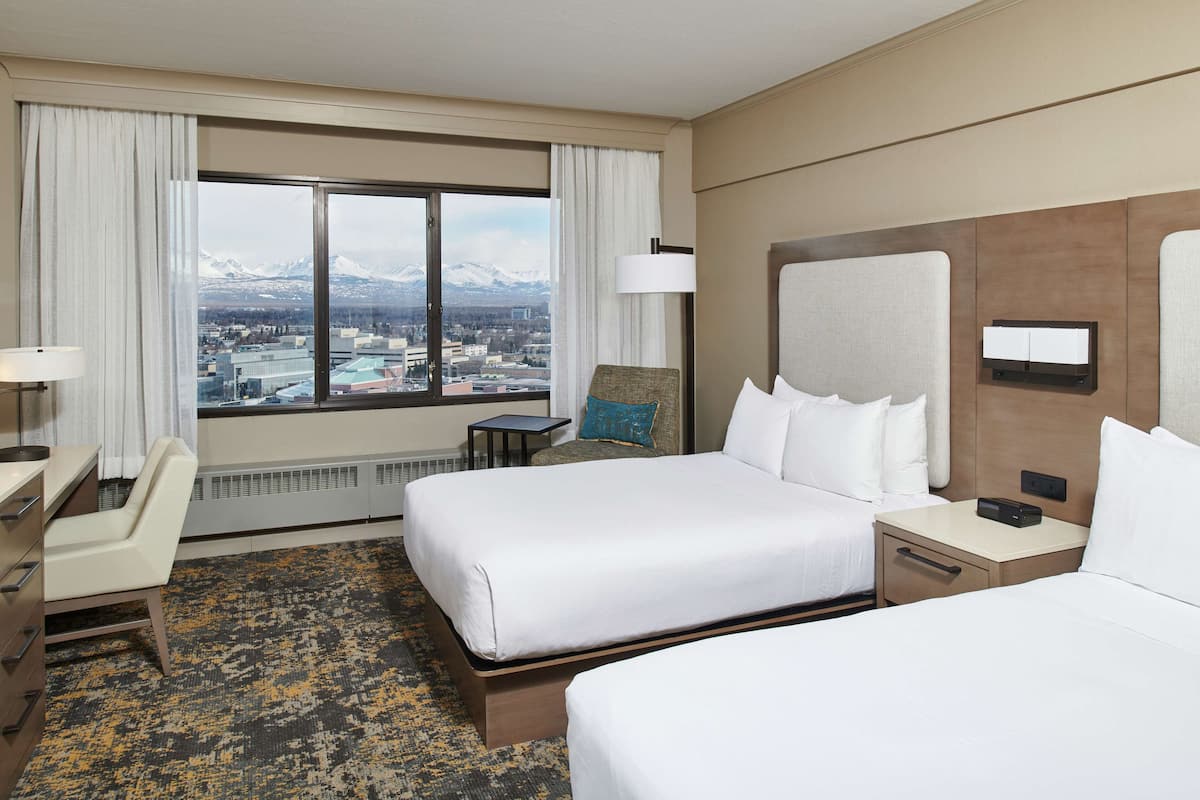 A hotel room with two beds, a desk, a chair, a view of mountains, and a large window.