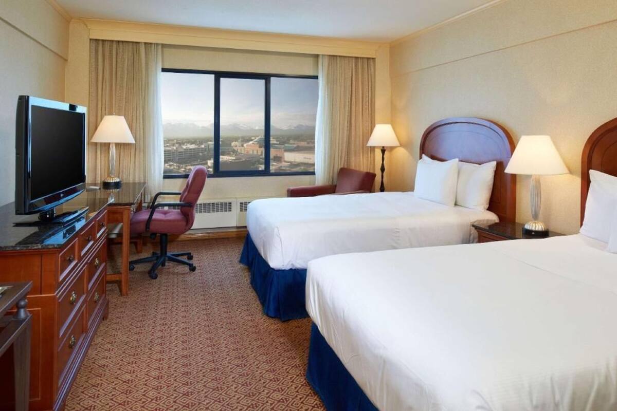 A hotel room with two beds, a TV, a desk, and a view of a cityscape.