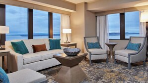 A hotel room with a sofa, armchair, and a small table with a bowl. Large windows offer a view of the ocean. - Hilton Anchorage (Anchorage)