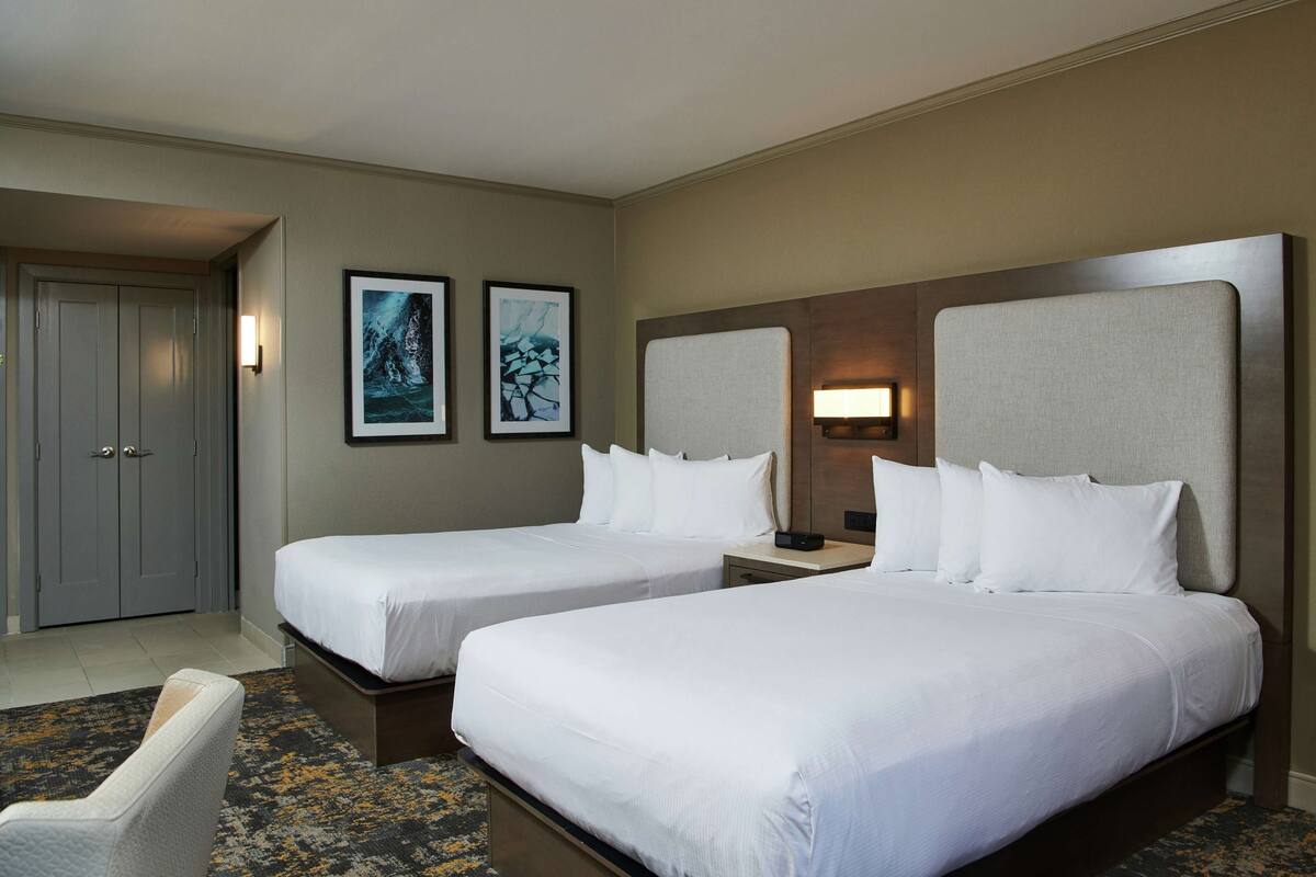 A hotel room with two single beds, a desk, a chair, and two framed pictures on the wall.