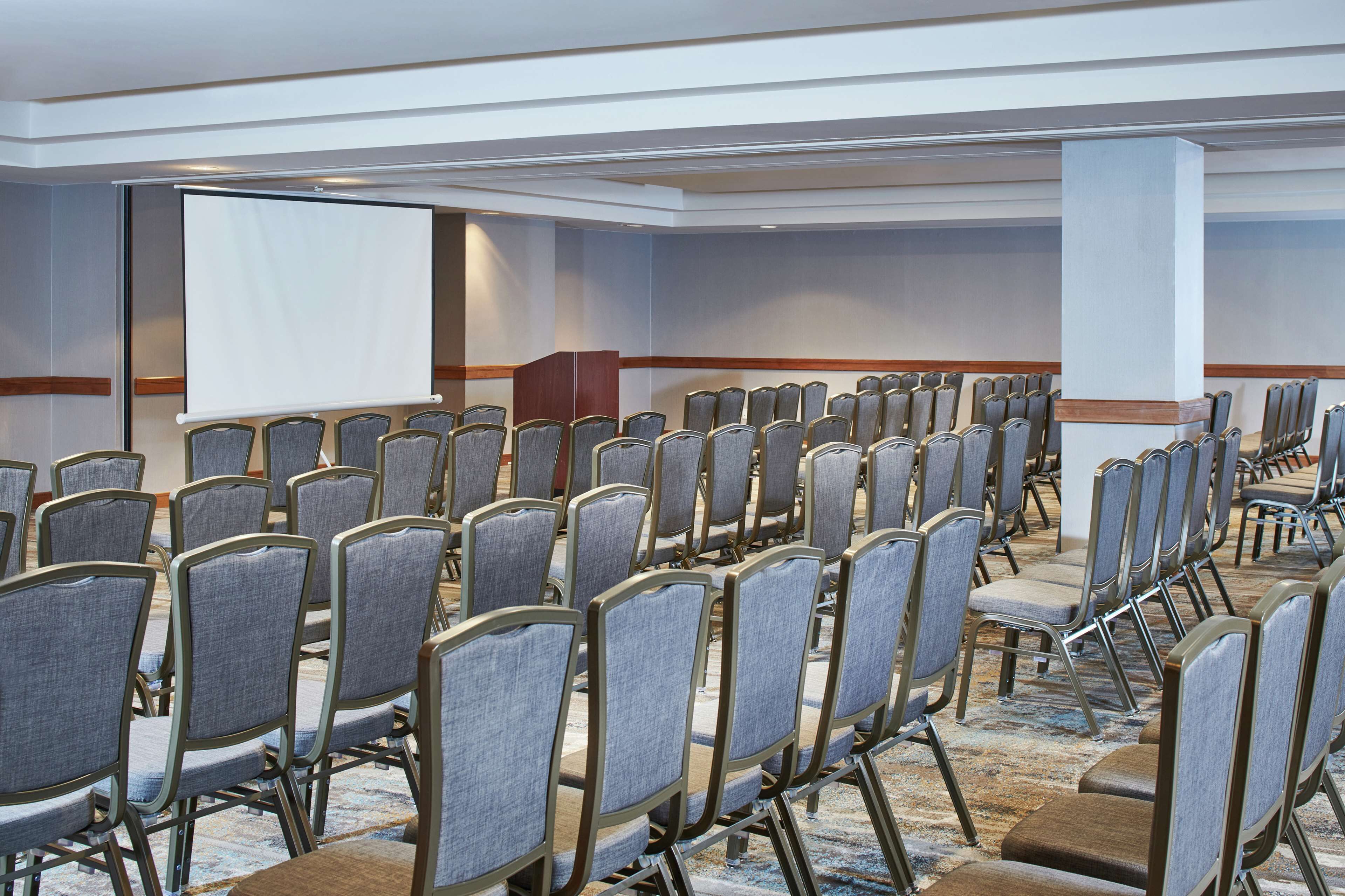 Un salón de conferencias con filas de sillas grises, una pantalla grande y un atril.
