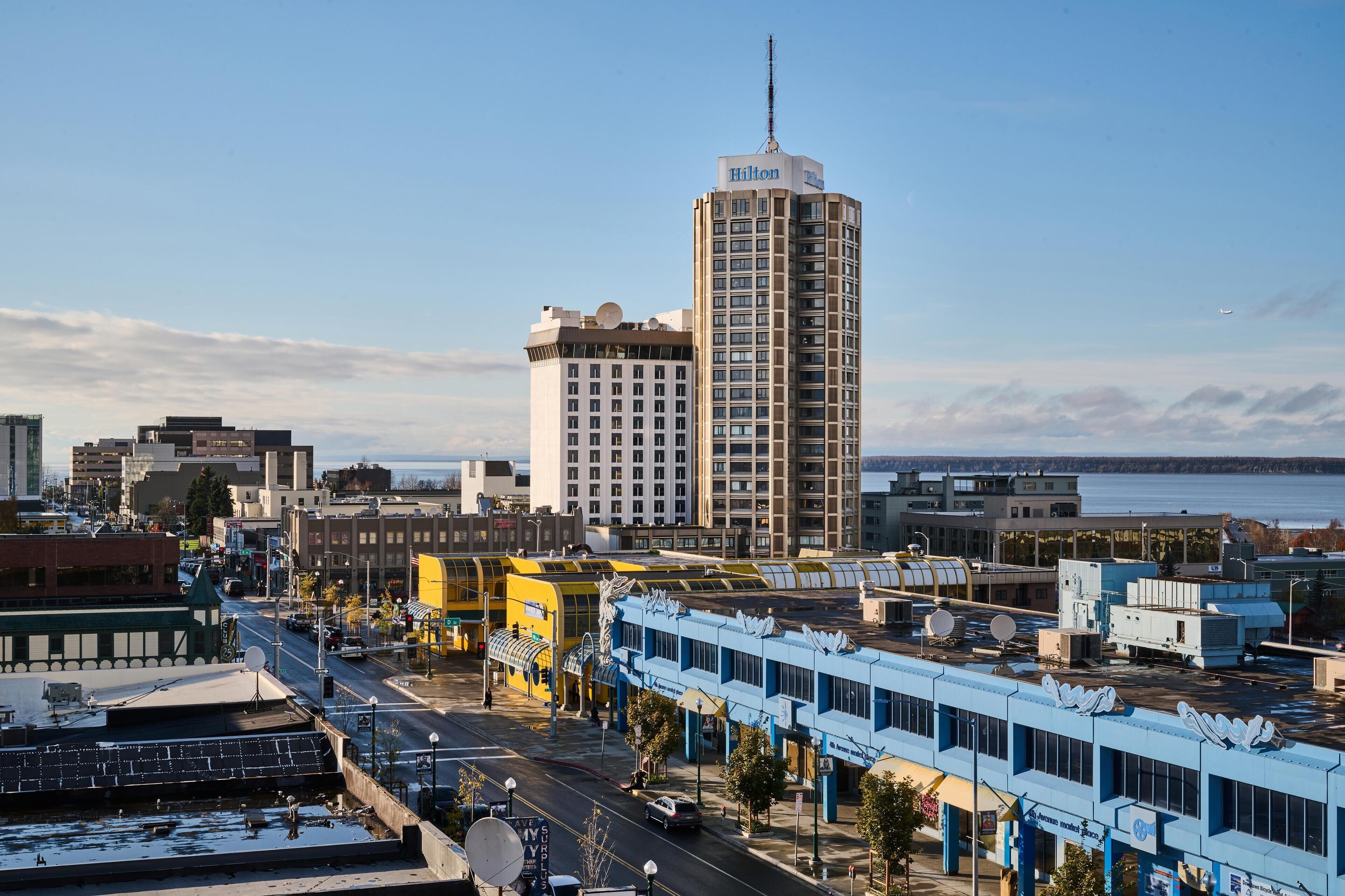 A tall hotel with a prominent antenna, surrounded by various buildings and a body of water in the background.