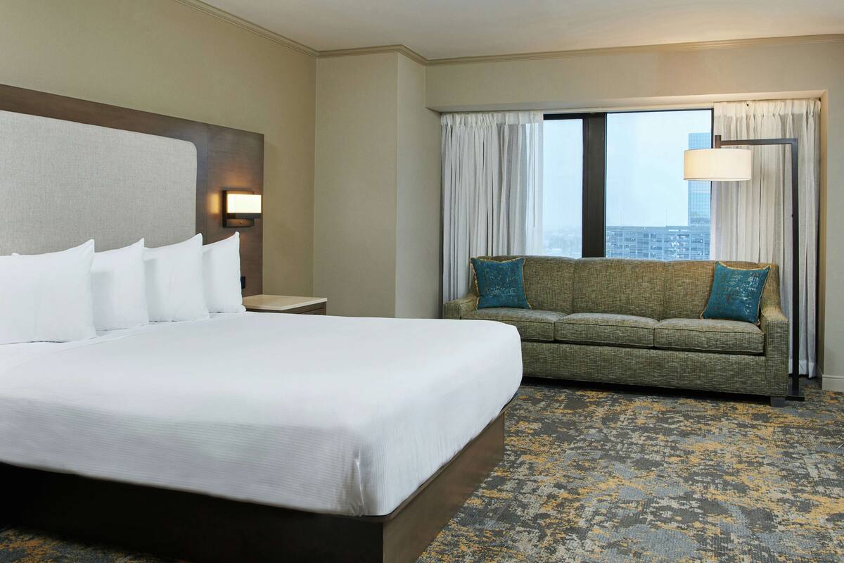 A hotel room with a large bed, a sofa, a lamp, and a view of the city through the window.